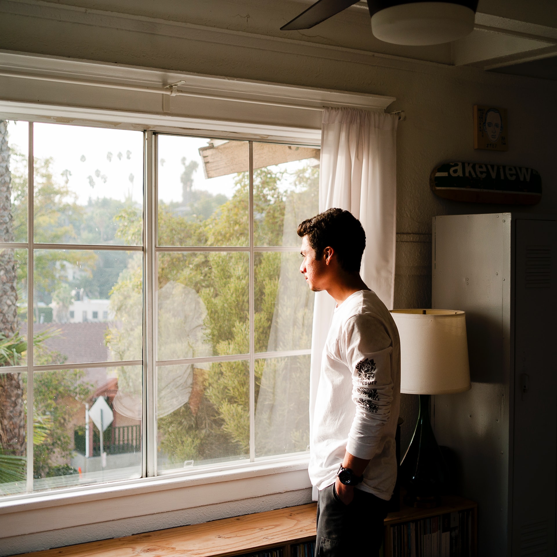 一个人站在一所房子里，凝视着一扇白色的大窗户，看着外面的街道。